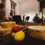 Na pierwszym planie znajdują się różne instrumenty muzyczne, na drugim - scena z muzykami.